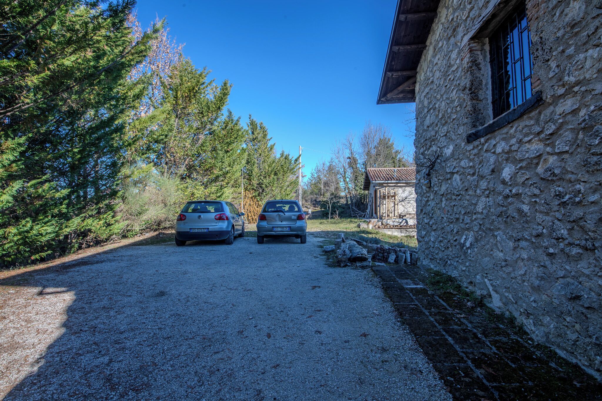 A gravel driveway with two parked cars, a stone building with a window, and a wooden structure in the background.
