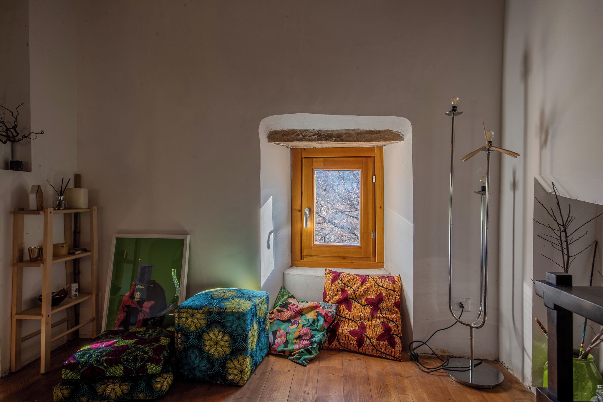 Um quarto aconchegante com piso de madeira, janela com vista para árvores, abajur e almofadas coloridas.
