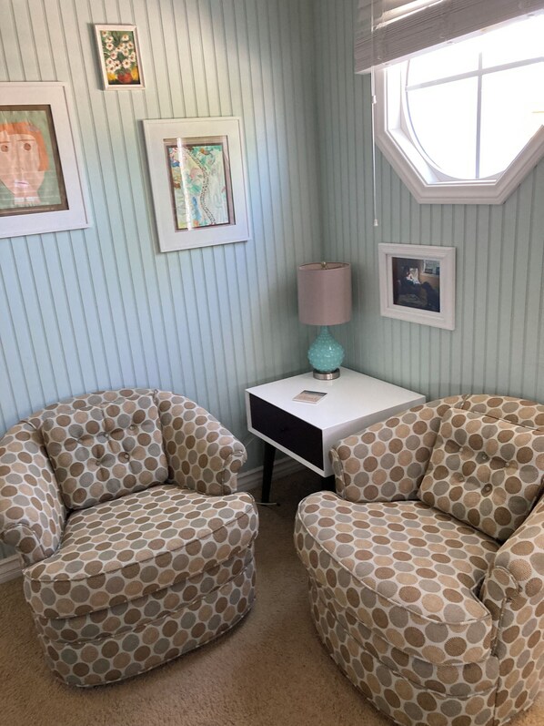 Une pièce comprenant deux fauteuils à motifs, une petite table avec une lampe et des œuvres d’art encadrées accrochées aux murs.