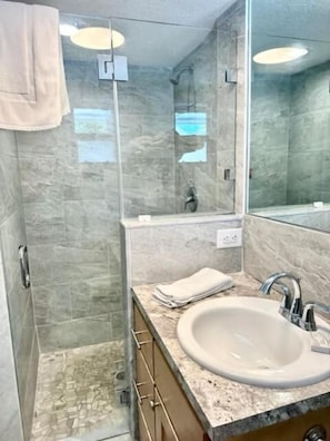 A modern bathroom with a glass shower enclosure, a marble-tiled shower area, a sink with a marble countertop, and a large mirror. - Matecumbe Resort (Islamorada)