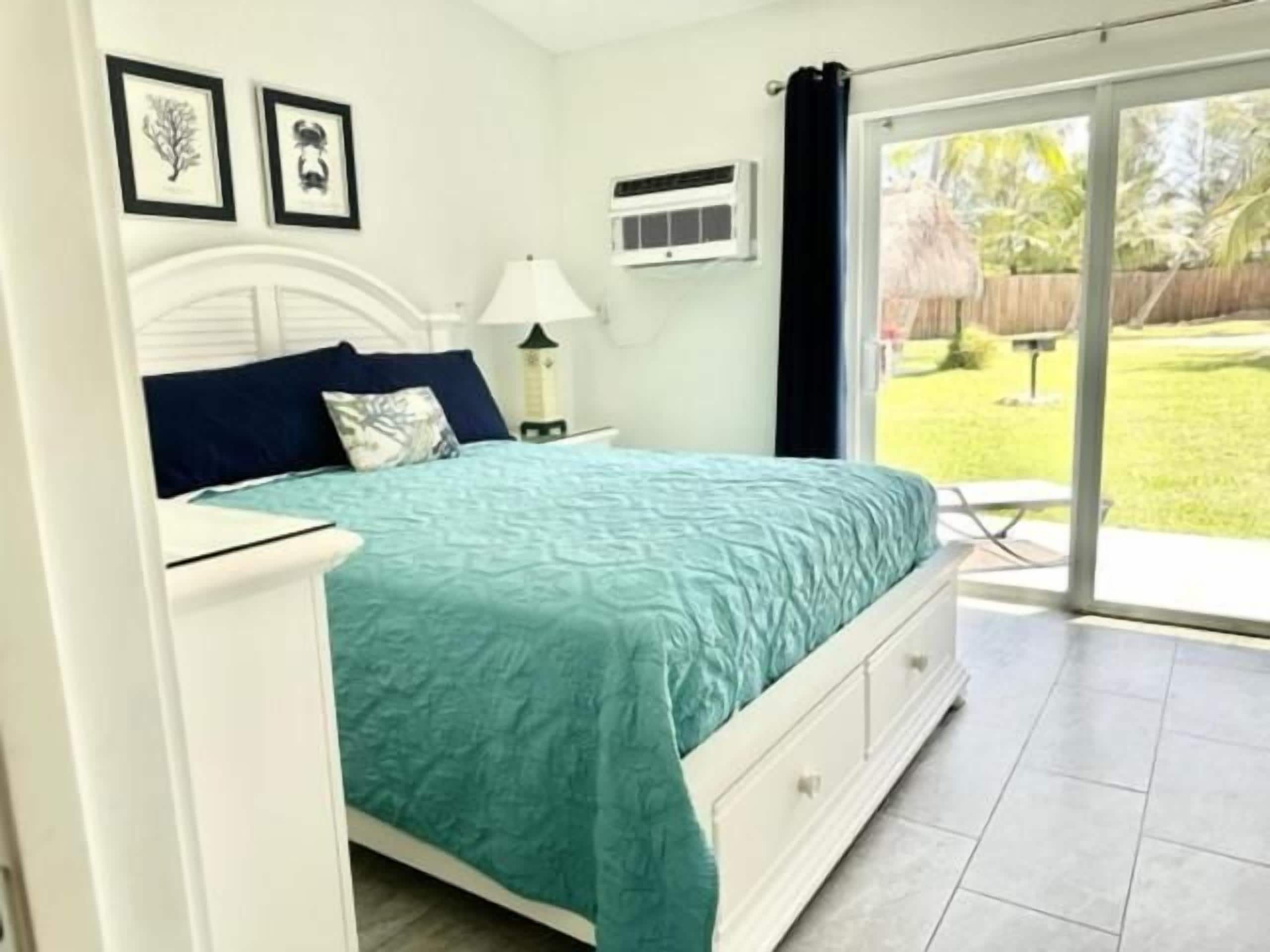A bedroom with a bed, a nightstand, a lamp, and a window with a view of a garden.