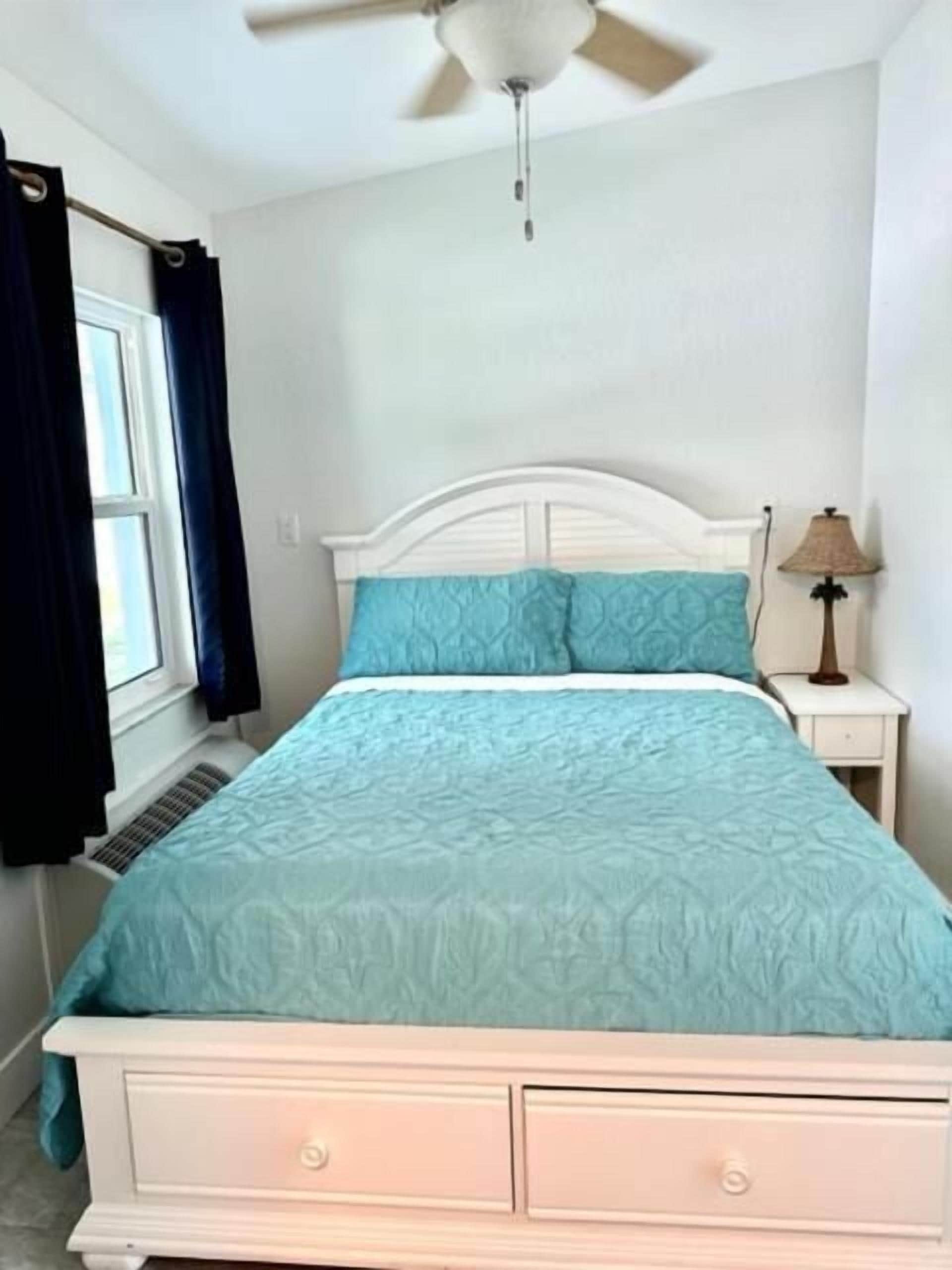 Une chambre à coucher avec un lit blanc, une literie de couleur turquoise, un ventilateur de plafond, une fenêtre avec des rideaux et une lampe de chevet.