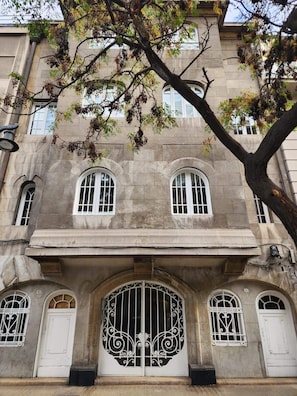 Un bâtiment historique doté d’une grande entrée en arc de cercle, de deux fenêtres ornées de grilles en fer décoratives et d’un arbre au premier plan.