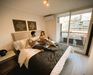 Ein Schlafzimmer mit einem Bett, zwei Kissen, einem Nachttisch und einem Fenster mit Blick auf Gebäude.