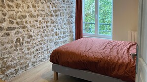 Une chambre avec un mur en pierre, un lit recouvert d’une couverture marron, un plancher en bois et une fenêtre donnant sur un paysage verdoyant.