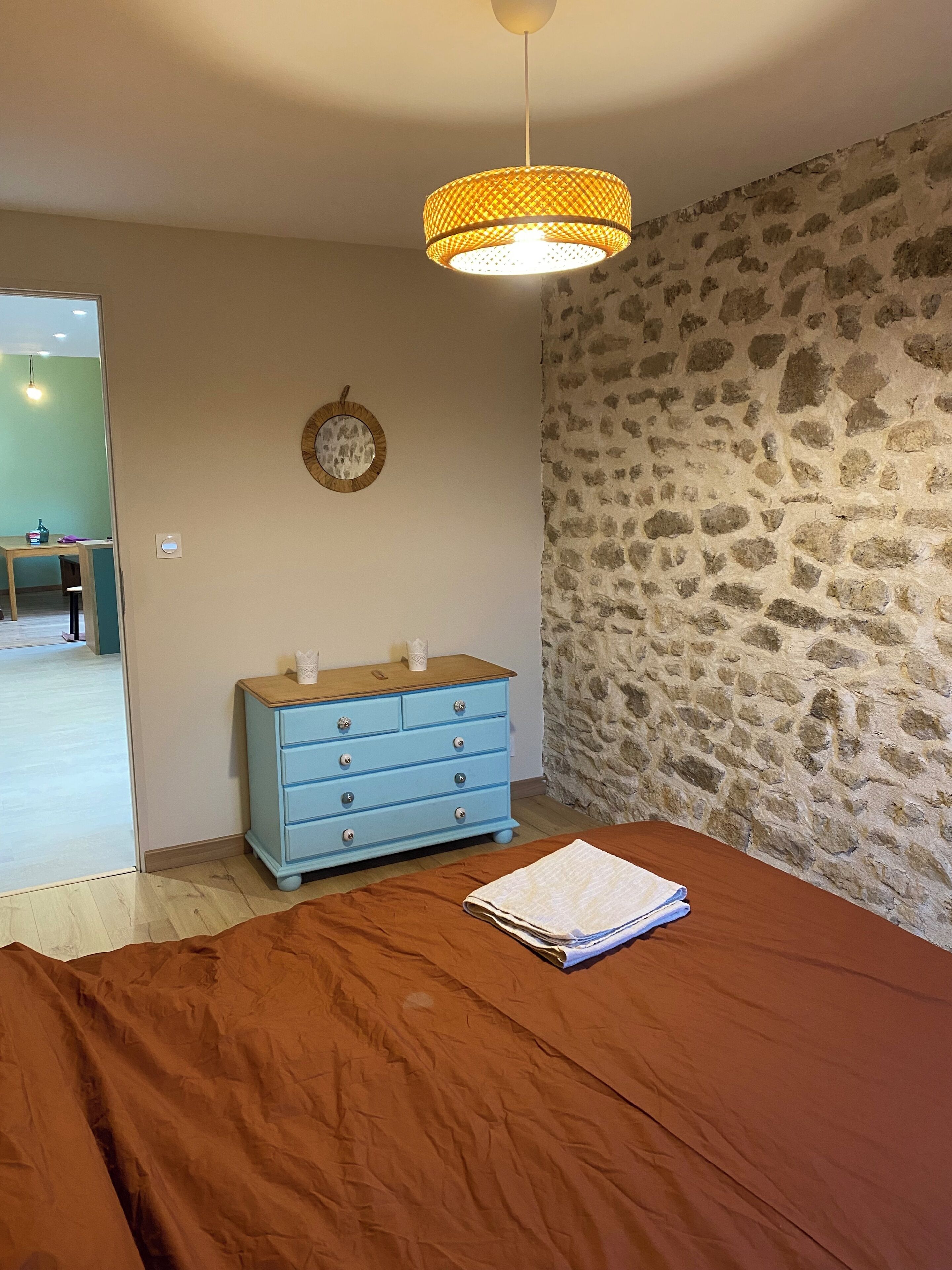 A bedroom with a stone wall, a blue dresser, a bed with a brown bedspread, and a hanging lamp.