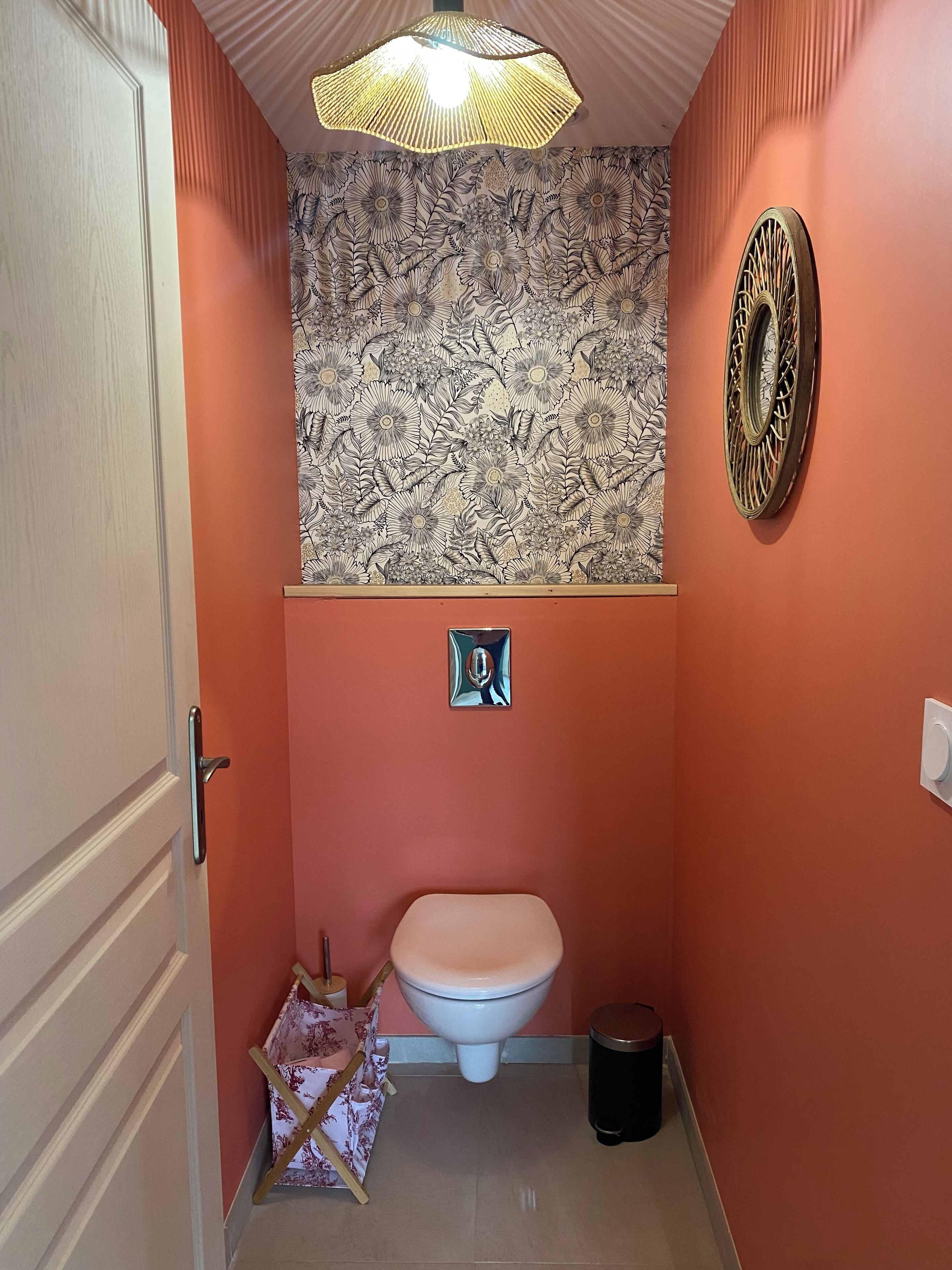 Une salle de bain comprenant des toilettes, une poubelle, un élément décoratif mural et un luminaire au plafond.