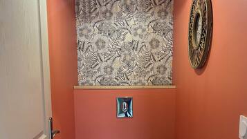 Une salle de bain comprenant des toilettes, une poubelle, un élément décoratif mural et un luminaire au plafond.