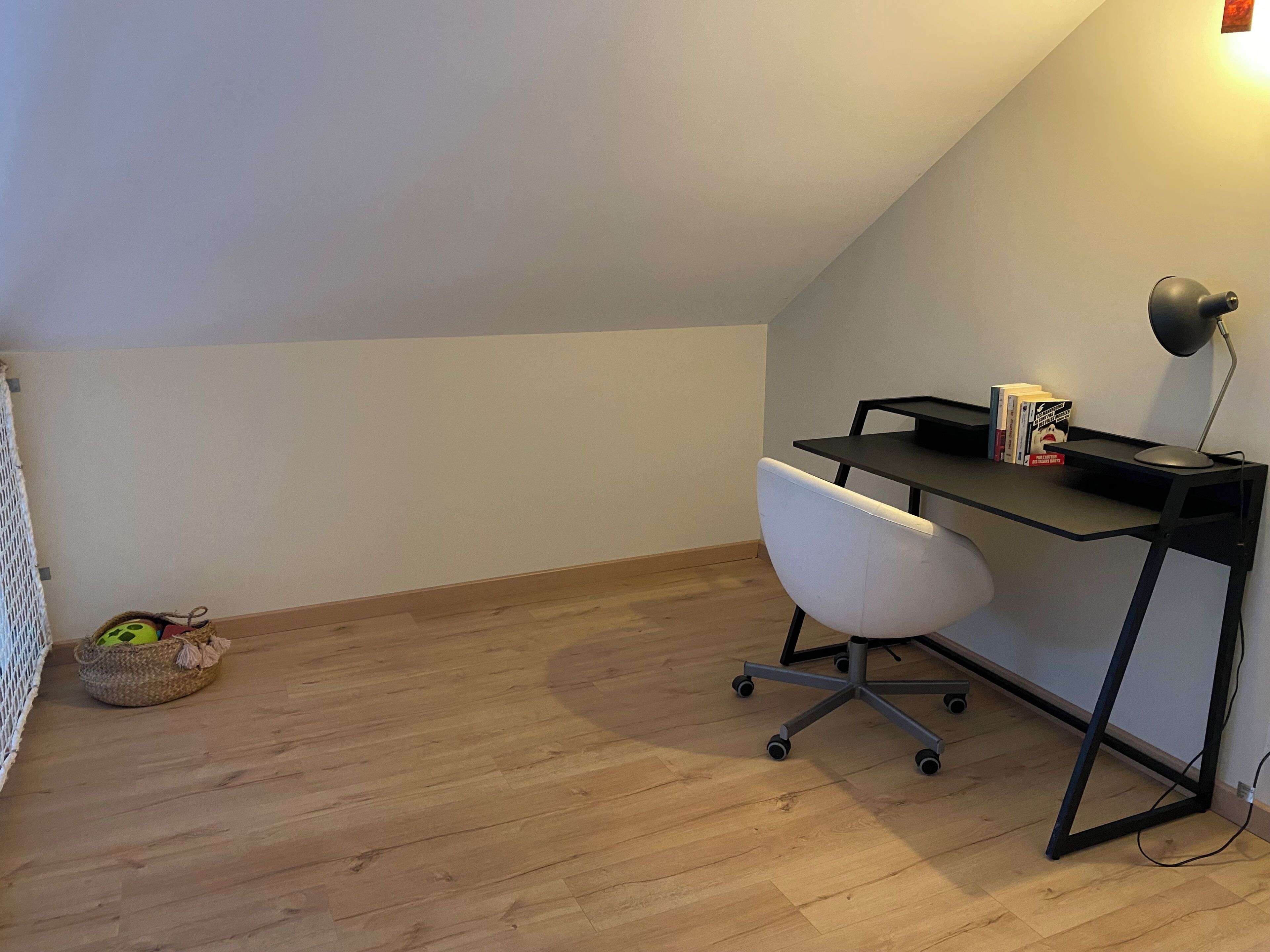 A small, sloped ceiling room with a wooden floor, a white office chair, a black desk with a lamp, and a radiator.