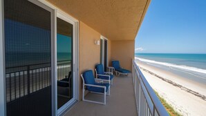 A balcony with a view of the beach, featuring a railing, a series of blue chairs, and a glass door. - Oceanfront 2BR Condo with Stunning Ocean Views and Beach Access, 505 (Indian Harbour Beach)