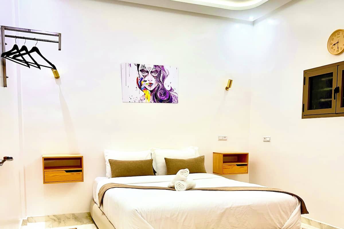 A modern bedroom with a large bed, a wall-mounted artwork, a clock, and a small cabinet.