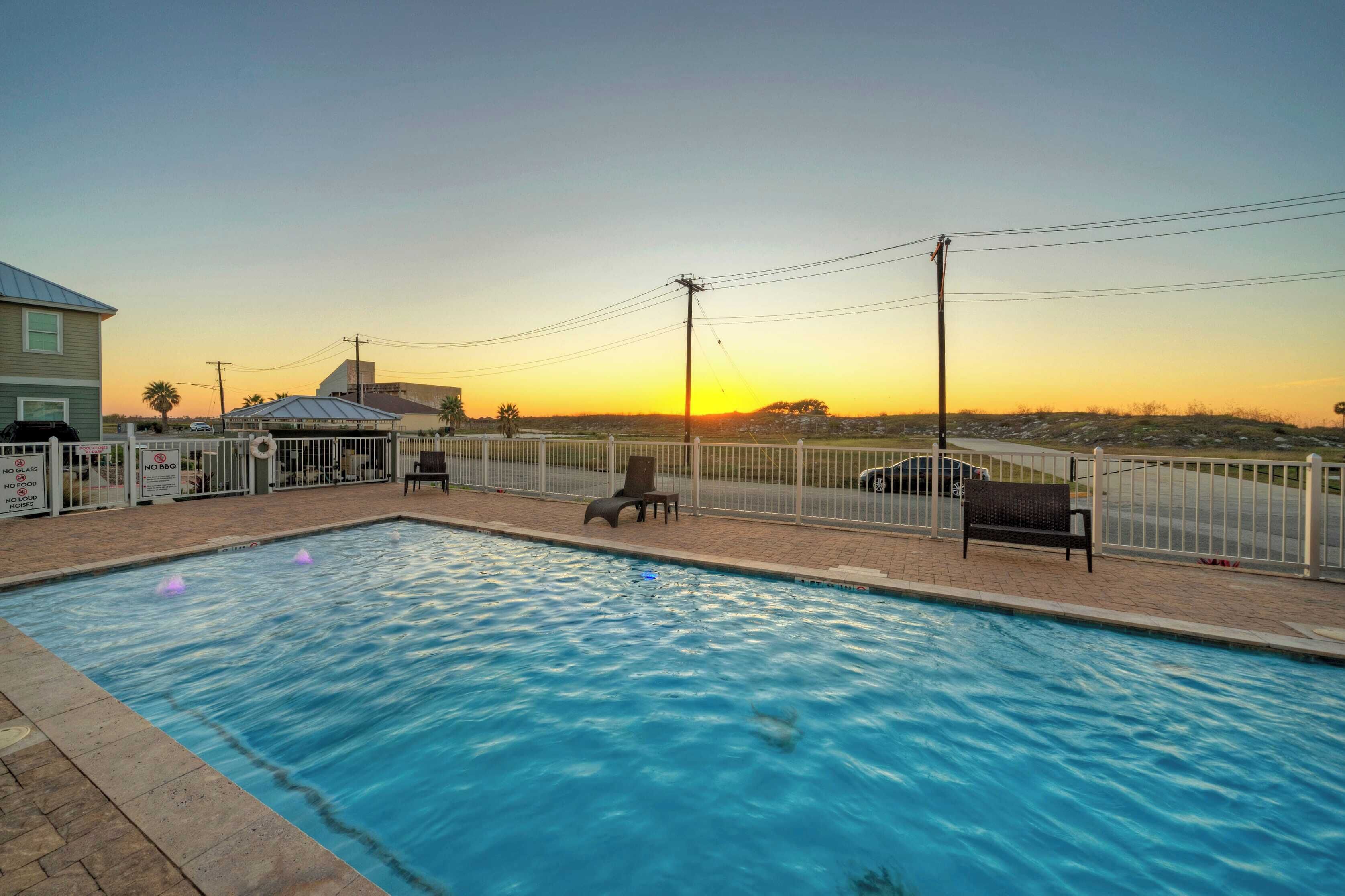 A swimming pool with a sunset view, surrounded by a fence, benches, and a covered area.
