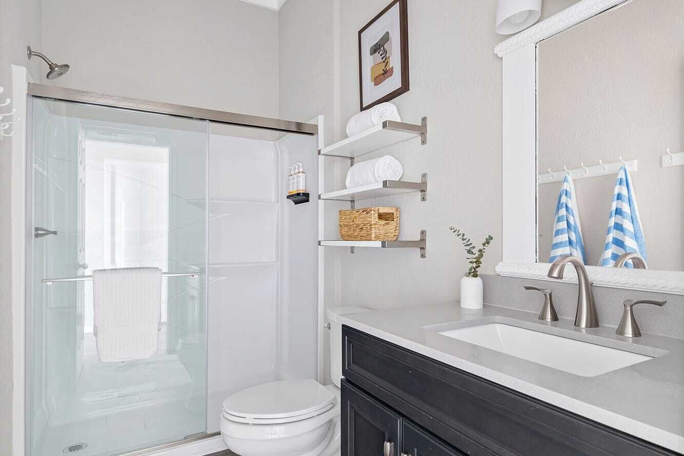 Un baño moderno con cabina de ducha de vidrio, inodoro blanco, tocador doble y espejo sobre el lavamanos.