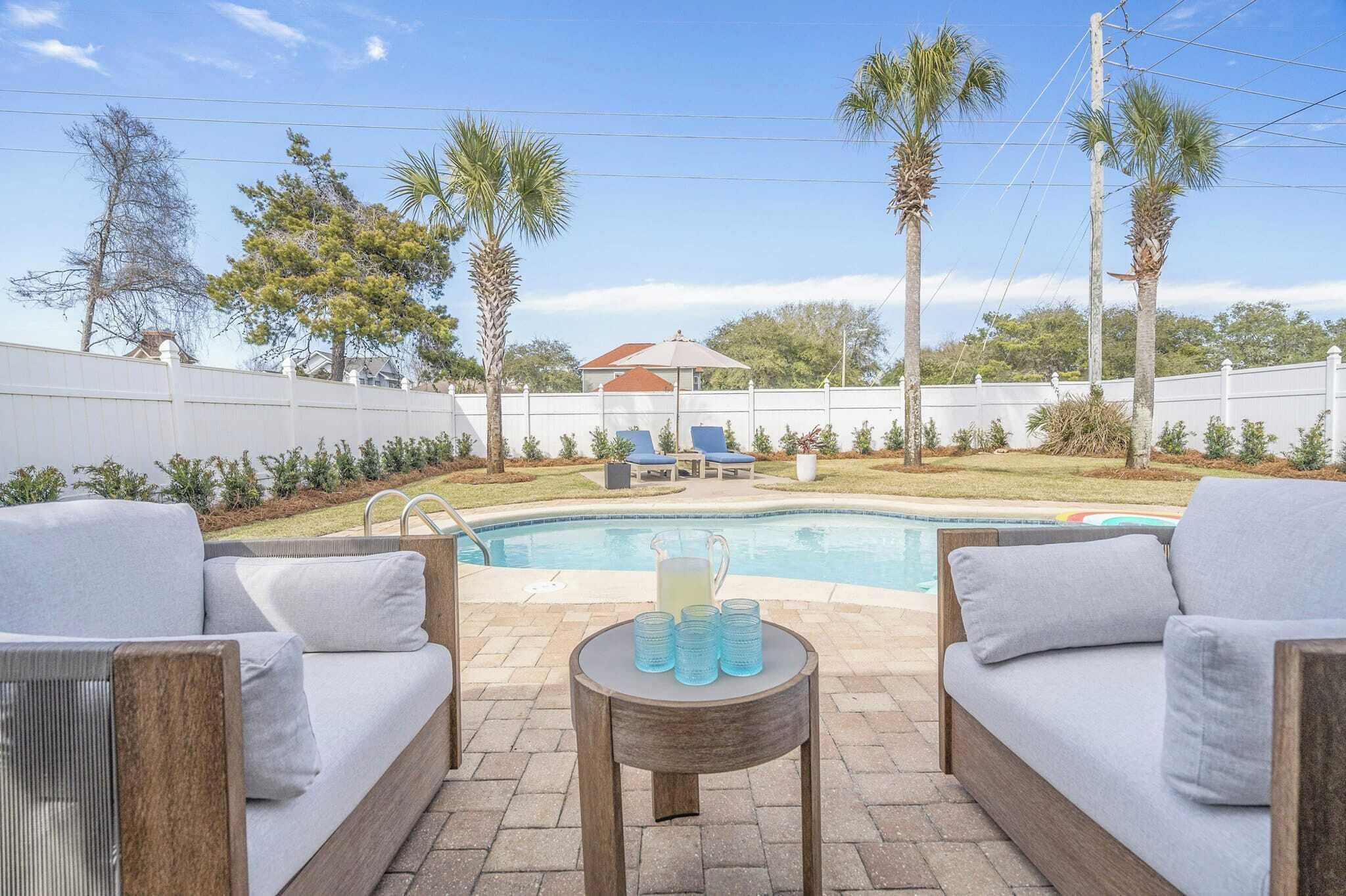 A backyard with a swimming pool, outdoor furniture, and palm trees.
