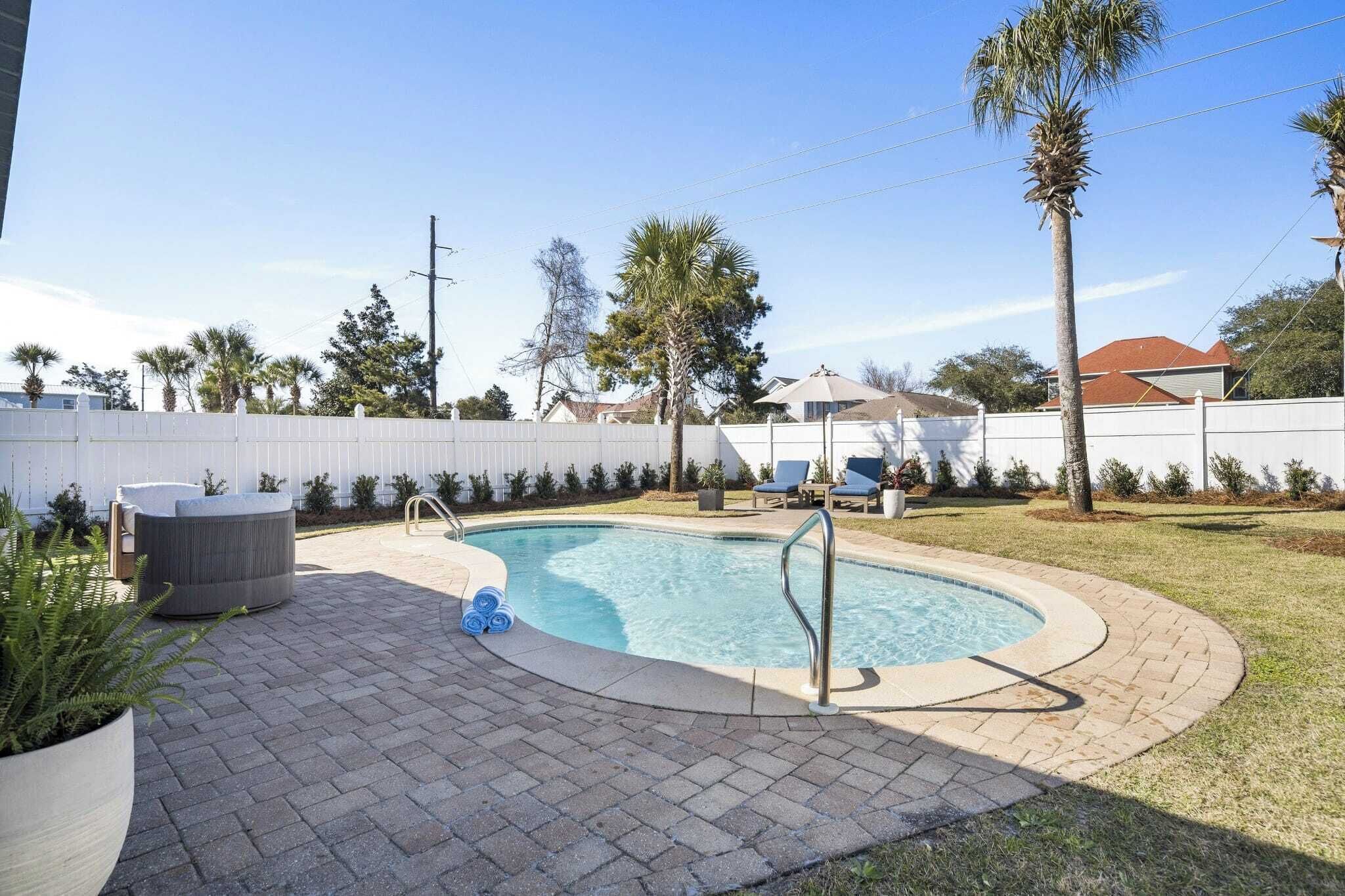 Una piscina con una escalera metálica, rodeada por un patio de ladrillo y una valla blanca.