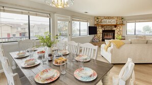 A dining area with a wooden table set for a meal, a stone fireplace with a 'BEACH' sign, and a beige sofa in the living room. - Mendel Manor by Avantstay Walk to Beach Large Deck Patio (Oceano)