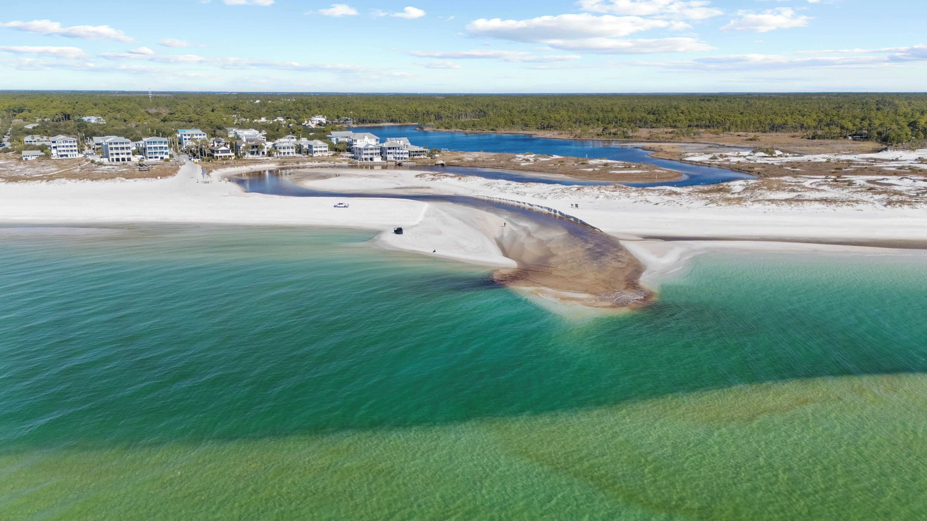 Vue aérienne d’une zone côtière avec une vaste plage de sable, une étendue d’eau d’un bleu limpide et un ensemble de bâtiments en arrière-plan.