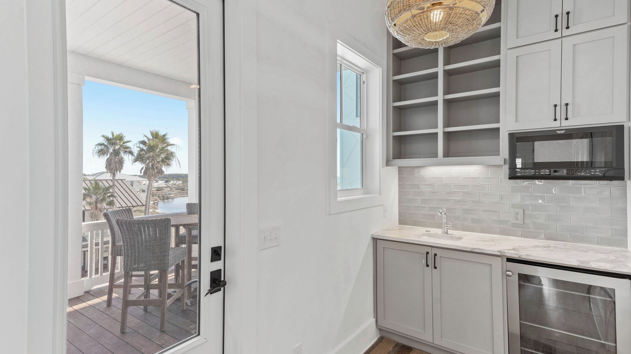 A kitchen with white cabinets, a microwave, and a window overlooking a deck with chairs.