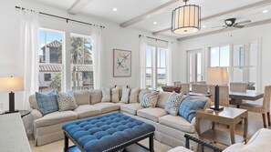 A spacious living room with a large sectional sofa, a blue ottoman, a dining area with wicker chairs, and large windows offering a view of the beach. - Grayt Digs by Avantstay Next to Beach, Oceanview (Santa Rosa Beach)