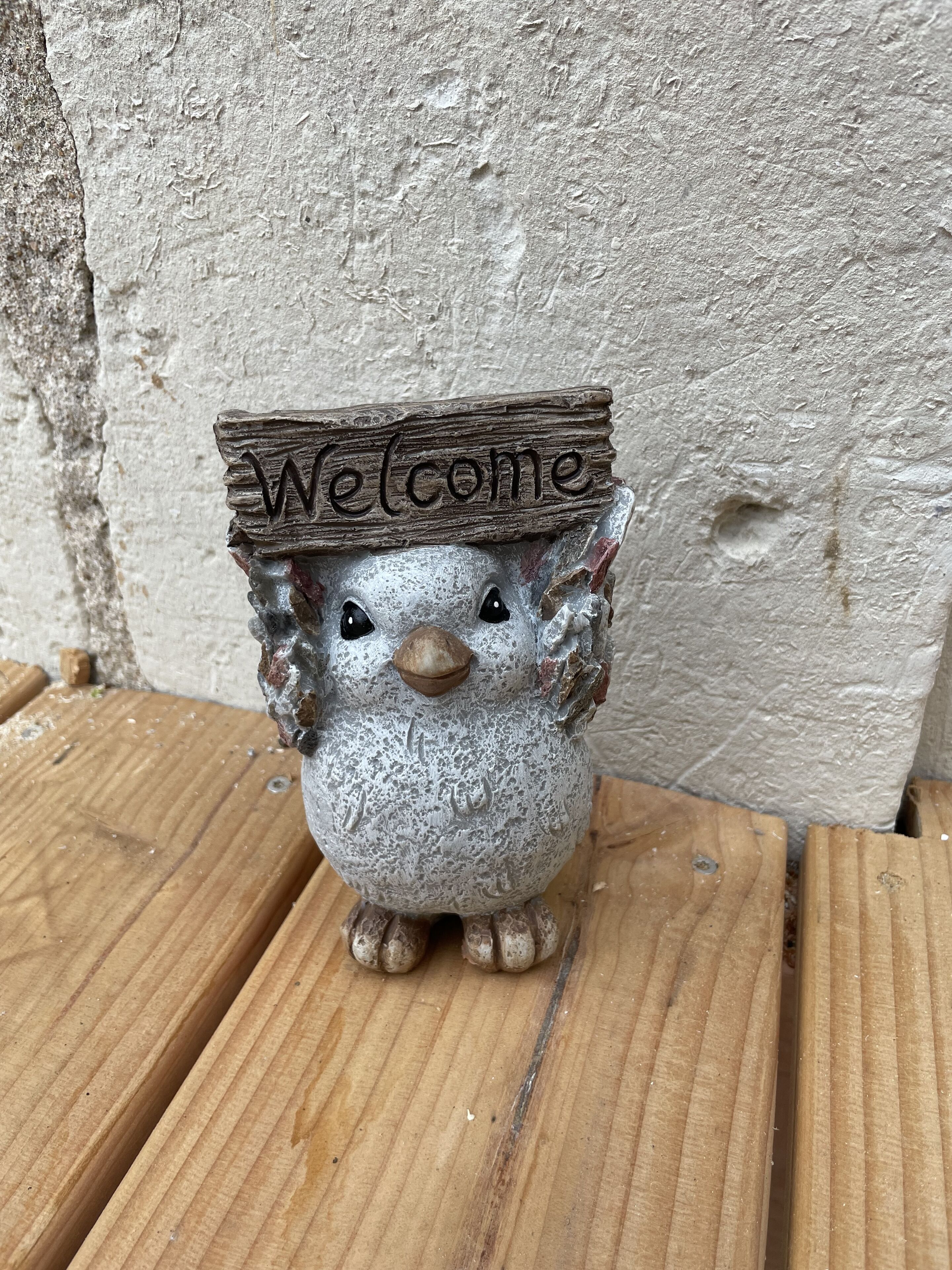 A small, rustic owl figurine with a 'Welcome' sign on its head, placed on a wooden surface against a textured wall.