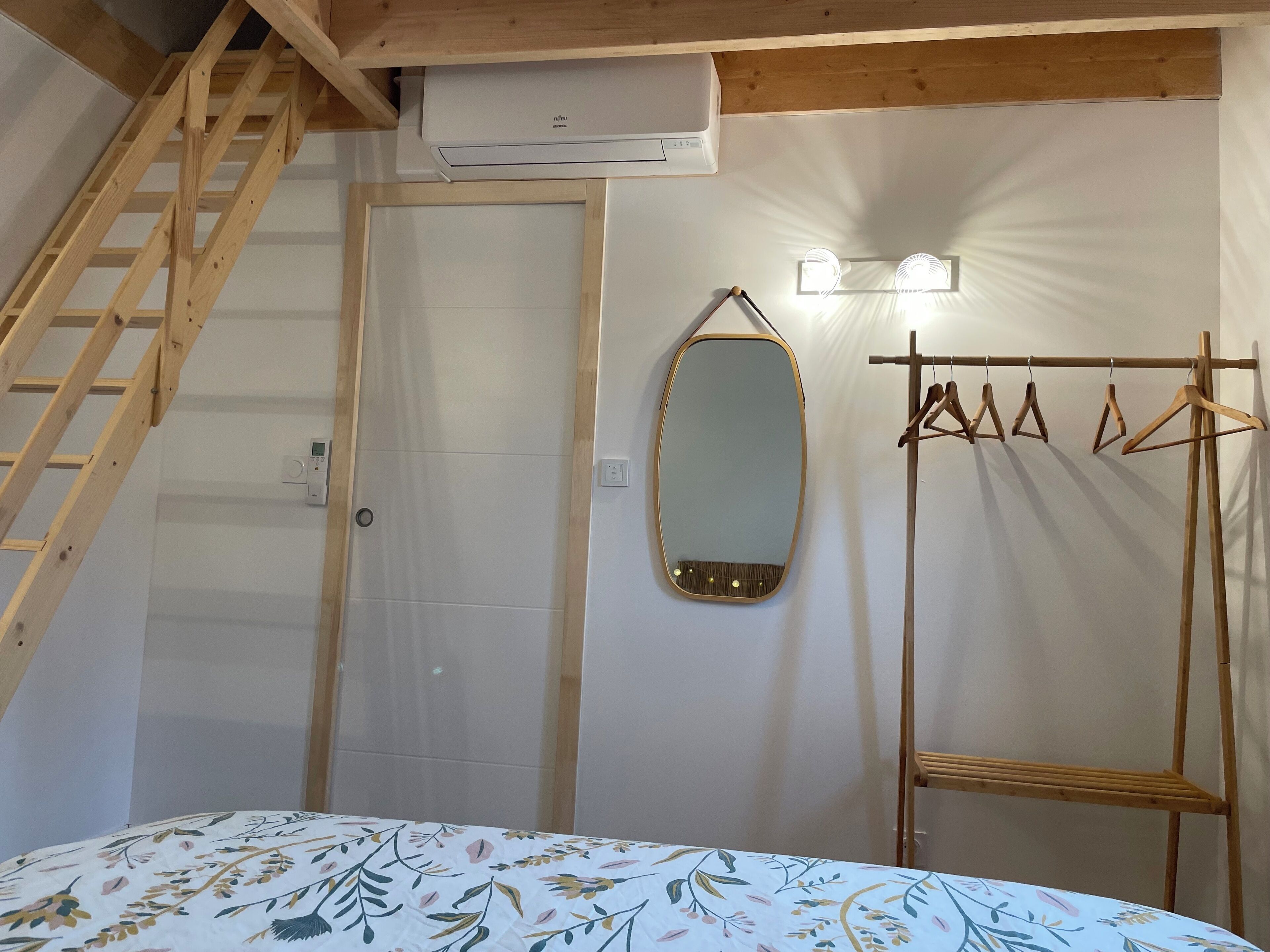 Un dormitorio con una escalera de madera, un equipo de aire acondicionado, un espejo y un perchero.