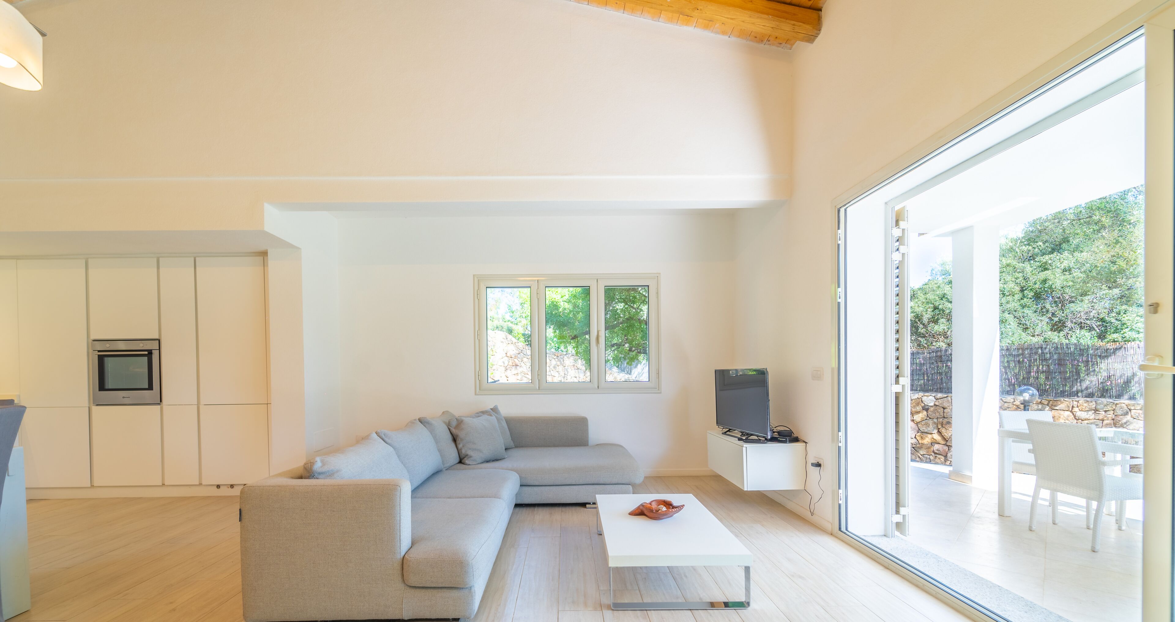 A modern living room with a grey sofa, a white coffee table, and a TV on a white stand. There is a large window providing a view of greenery outside.