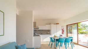 Moderni keittiö, jossa on valkoiset kaapit ja laatoitettu seinäosa työtason yläpuolella, ruokailutila sinisillä tuoleilla sekä suuri ikkuna, josta avautuu näkymä ulos.