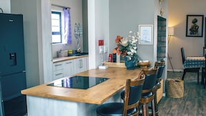 A modern kitchen with a wooden island, bar stools, a refrigerator, and a dining area with a table and chairs. - Spacious 3BR Family Haven in Walvis Bay – 30 Min to Swakopmund! Self-Catering (Walvis  Bay)