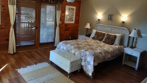 A bedroom with a large bed, a ceiling fan, wooden walls, a window with blinds, and two bedside lamps. - Tropical Room at Cabin on the Lake (Lake Helen)