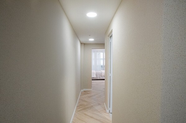 A narrow hallway with a wooden floor and beige walls, leading to a room with a bed and a window.