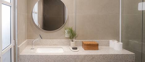 A modern bathroom with a granite countertop, a round mirror, a small potted plant, a wooden box, and two rolls of toilet paper.
