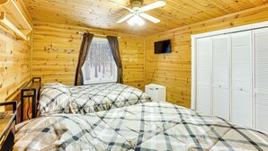 Habitación de cabaña de madera con dos camas, un ventilador de techo, un televisor y una ventana con cortinas.