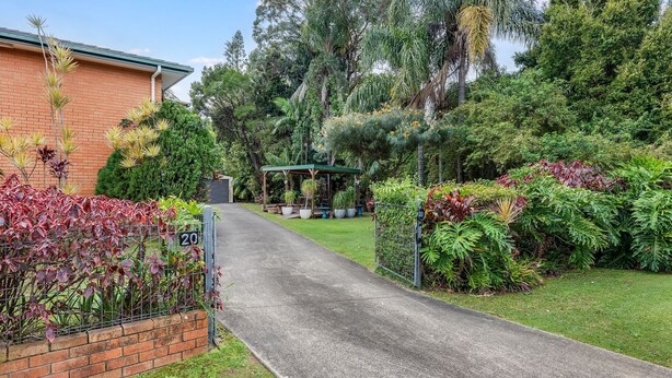 A residential driveway leading to a house with a brick facade, surrounded by lush greenery and a small gazebo.