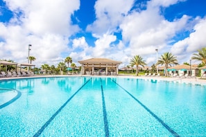 Une grande piscine extérieure à l’eau d’un bleu limpide, entourée de chaises longues et de parasols, et un pavillon couvert en arrière-plan.