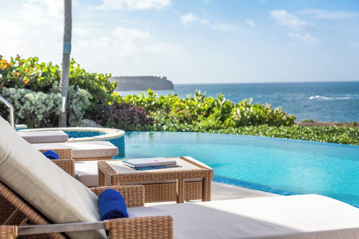 Ein Poolbereich mit Korbstühlen, einem kleinen Tisch und einem Buch, mit Blick aufs Meer.