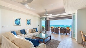 A spacious living room with a large glass coffee table, a beige sofa with blue cushions, and a view of the ocean through an open door.