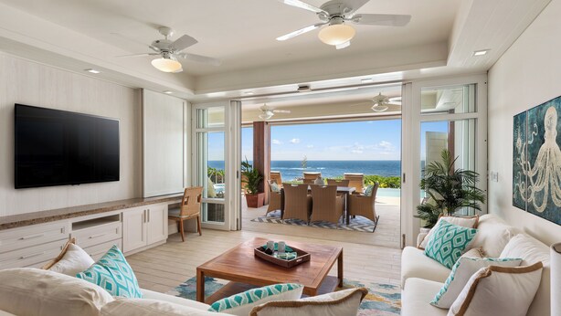 A spacious living room with a flat-screen TV, comfortable seating, a coffee table, and a view of the ocean through large sliding glass doors.