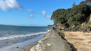 Une promenade en béton le long d’une plage, avec des vagues qui se brisent sur le rivage, un ciel dégagé et une colline boisée en arrière-plan.