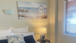 Un lit bien fait, agrémenté d’un coussin à motifs floraux bleus et blancs, d’une couverture bleu foncé et d’une serviette blanche. Une lampe est posée sur une table de chevet, et un tableau représentant une scène de plage est accroché au mur.