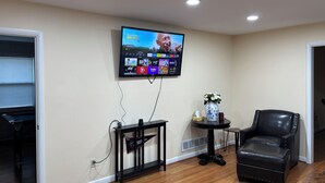 Ein Wohnzimmer mit an der Wand montiertem Fernseher, der eine Streaming-Dienstanwendung zeigt, einem kleinen Beistelltisch mit einer Vase, einem schwarzen Ledersessel und einem Holzboden.