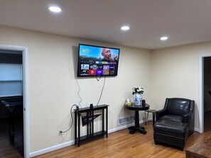 Un soggiorno con una TV a parete che mostra l'interfaccia di un servizio di streaming, un tavolino con un vaso, una poltrona in pelle nera e un pavimento in legno.