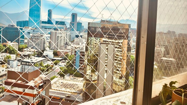 A cityscape viewed through a chain-link fence, featuring a mix of modern and older buildings, including a prominent blue skyscraper. - Vista al Costanera (Santiago)
