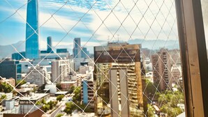 A cityscape through a chain-link fence with a prominent skyscraper in the background. - Vista al Costanera (Santiago)