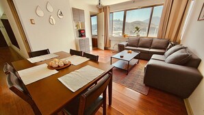 A modern living room with a wooden dining table, a sofa, and a coffee table. There is a kitchen area with a refrigerator and a water dispenser. The room has large windows with a view of the outdoors. - Vista al Costanera (Santiago)