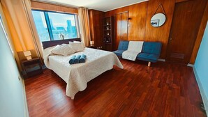 A hotel room with a large bed, a blue sofa, wooden paneling, a view of the city, and a circular mirror on the wall. - Vista al Costanera (Santiago)