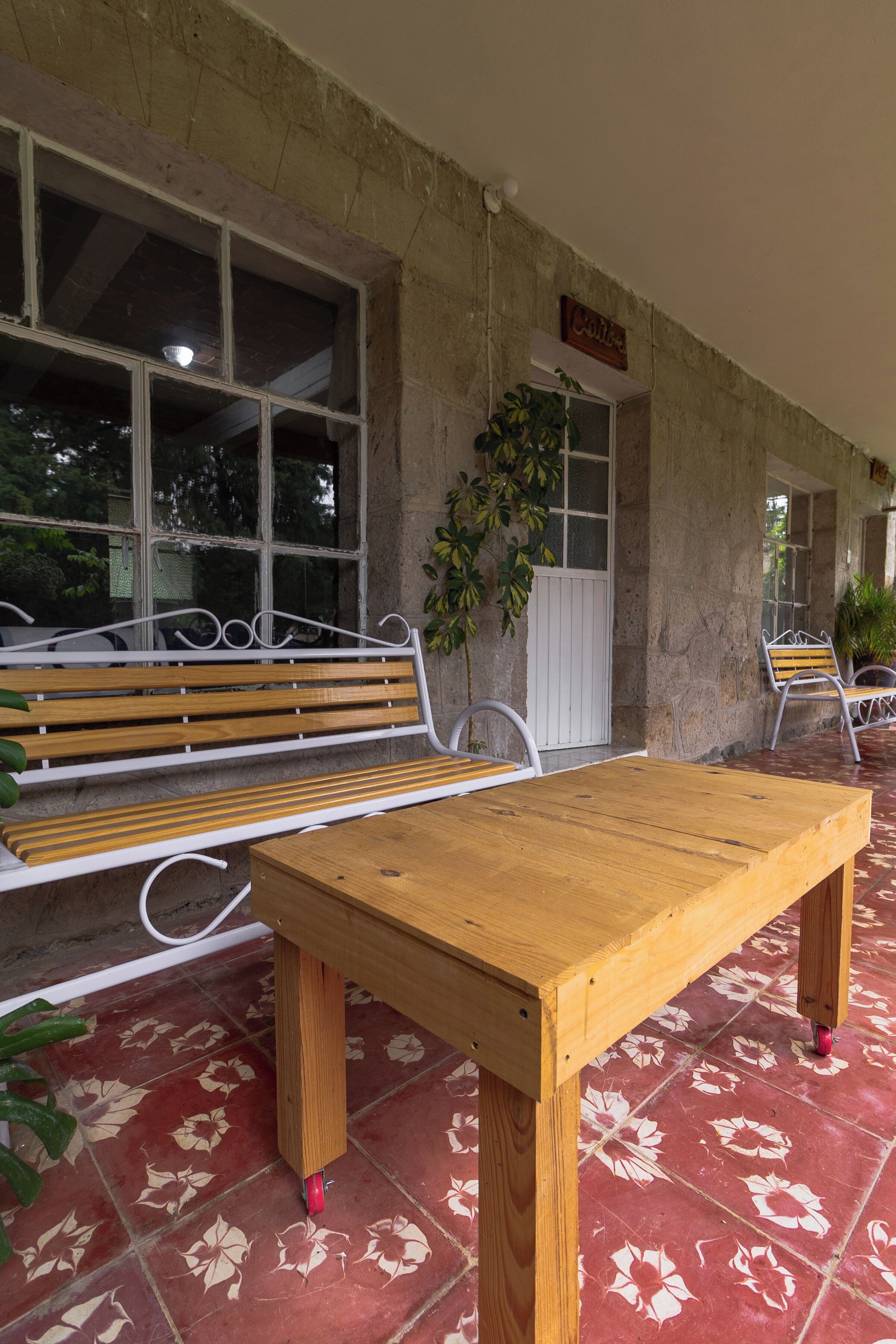 Un banco de madera y una mesa en un área exterior cubierta, con un muro de piedra y una ventana.