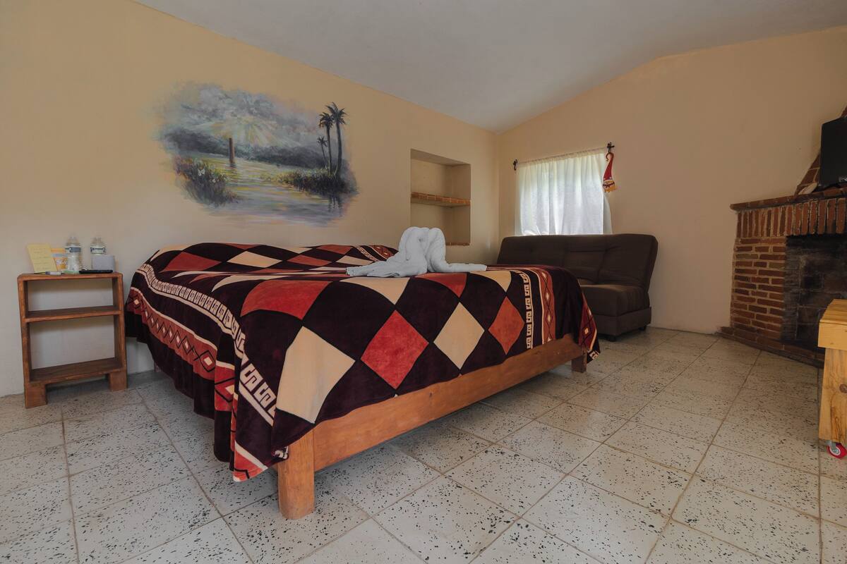 Un dormitorio con una cama, una silla, una mesita, un cuadro y una chimenea.