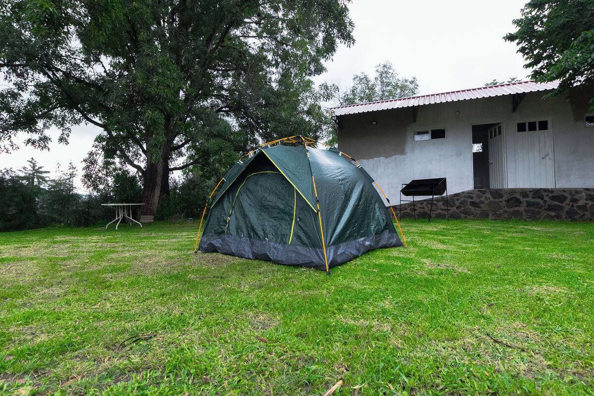 Una tienda de campaña verde montada sobre un césped frente a un edificio de base de piedra y techo rojo.