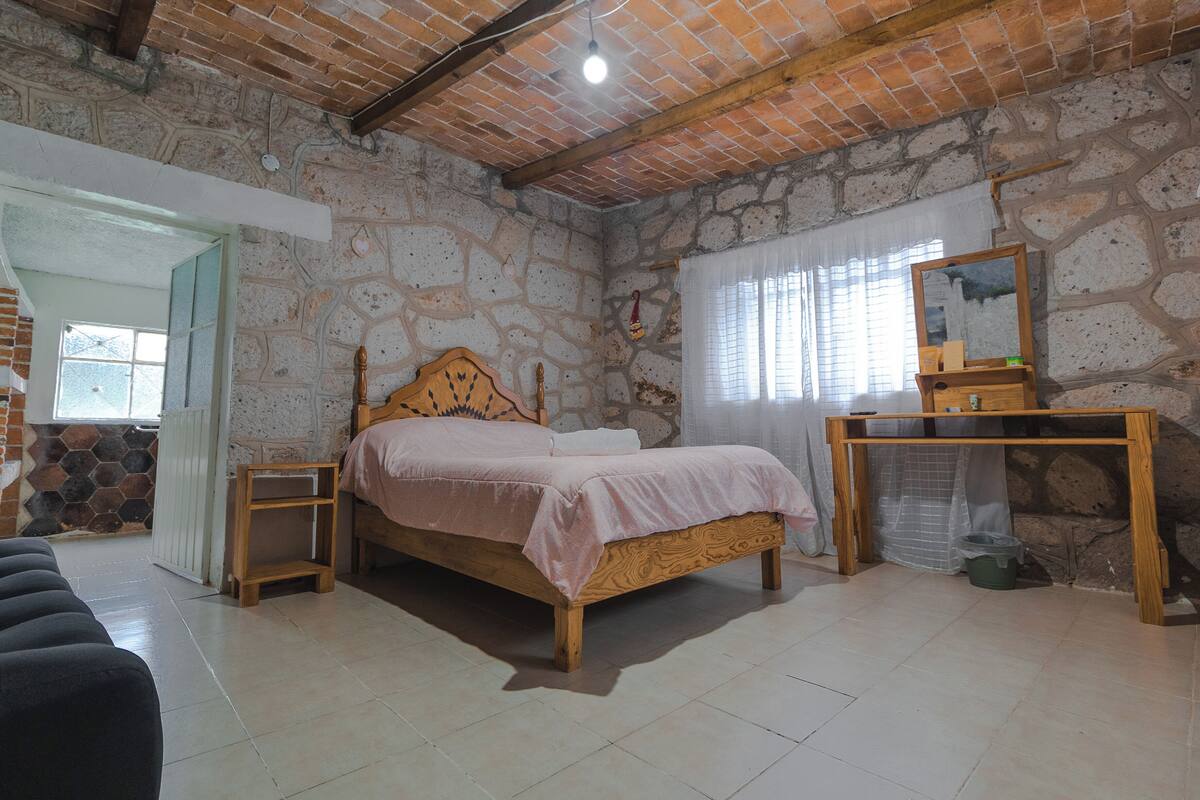 Un dormitorio con pared de piedra, cama de madera, mesita de noche, espejo y ventana con cortinas.