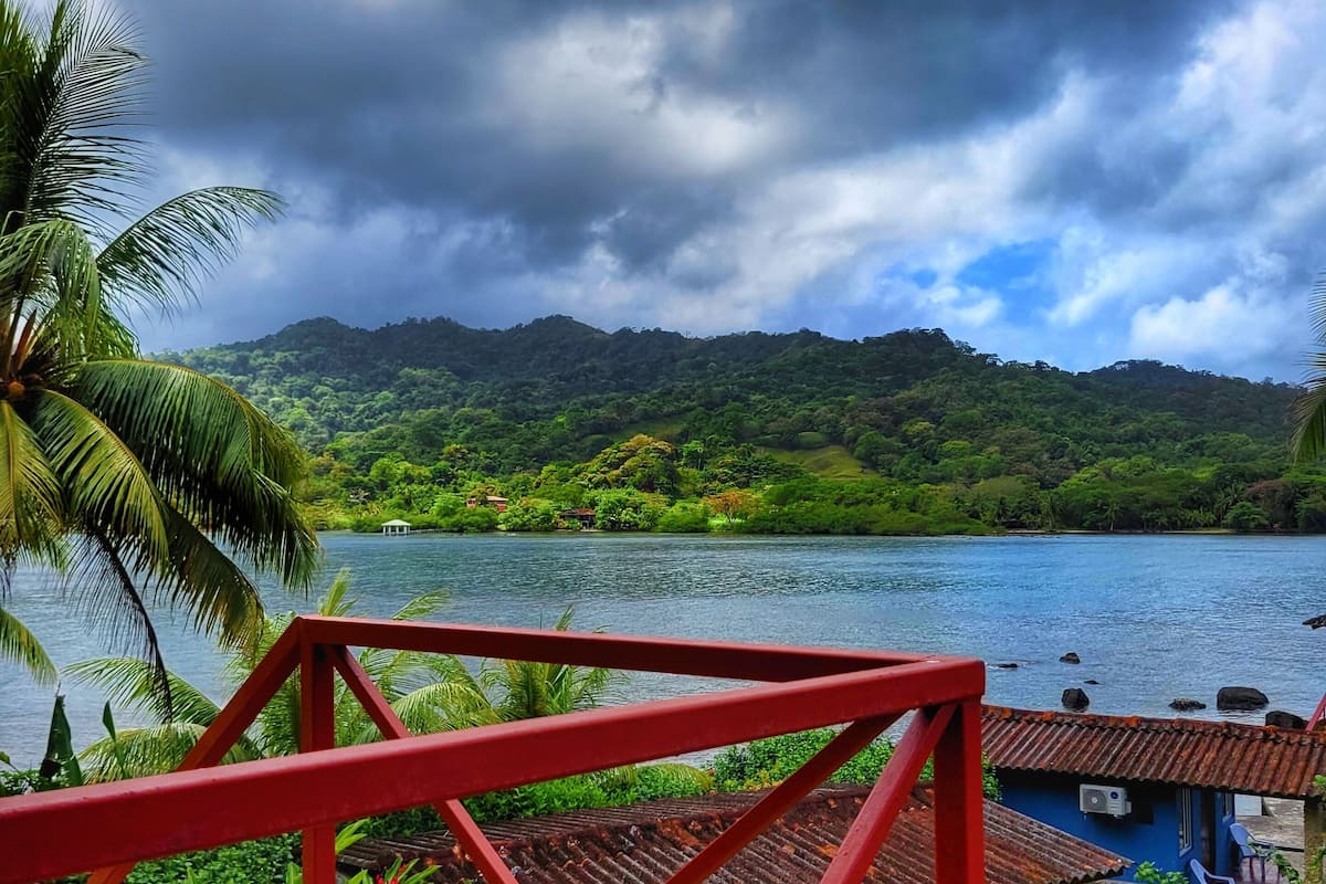 A view from a red metal railing overlooking a lake, with lush greenery and a mountain in the background.